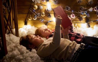 dve deti lezia na hunatej podlozke, citaju knihu a za nimi svietia vianocne svetla