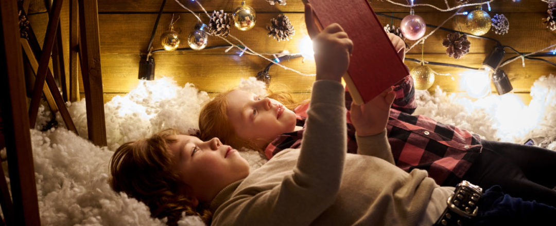 dve deti lezia na hunatej podlozke, citaju knihu a za nimi svietia vianocne svetla