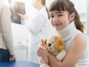Cute girl with cervical collar in the doctor's office: she is smiling, holding her teddy bear and giving a thumbs up