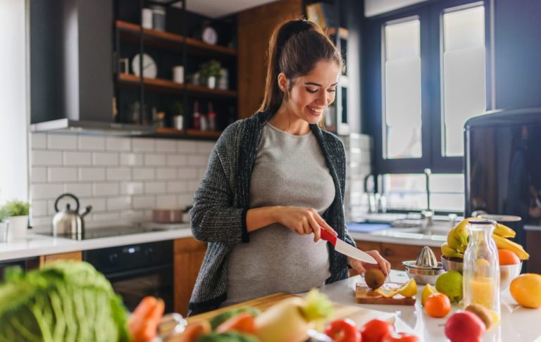Beautiful smiling young pregnant woman preparing healthy food with lots of fruit and vegetables at home kitchen strava v tehotenstve